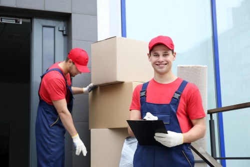 Two movers from St Johns Wood Man with Van carrying out house removal tasks outside a modern residential building. The mover in the foreground, wearing a red cap and a red T-shirt with navy blue overalls, is smiling at the camera and holding a clipboard. The second mover, also dressed in a red cap, red T-shirt, and navy overalls, is focused on stacking or adjusting large cardboard boxes, which are positioned on a small ramp or platform near the entrance. The background features a sleek, dark-colored door and a large glass window framed with blue trim, suggesting a contemporary residential or office property in the St Johns Wood area. The scene emphasizes professional and organized packing and loading activities typical of local removals services, with clear attention to safety and efficiency in house moving procedures.