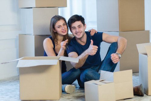 A young couple sitting on the floor of a bright, spacious room surrounded by open cardboard boxes and packing materials, indicating a house relocation process. The woman has long brown hair and is wearing a sleeveless navy top, while the man has short dark hair and is dressed in a dark t-shirt and jeans. They are smiling and appear relaxed, with the woman resting her arm on the man's shoulder and the man giving a thumbs-up gesture. In the background, there are stacked cardboard boxes of various sizes, some sealed and others open, revealing household items or packing supplies. The room is well-lit with natural light coming through large windows, and the setting suggests a residential area possibly within St Johns Wood, London, where local professional removals services like those offered by St Johns Wood Man with Van are commonly used for house moves. The overall scene captures the beginning stages of a move, emphasizing organized packing and teamwork in a typical UK suburban environment.
