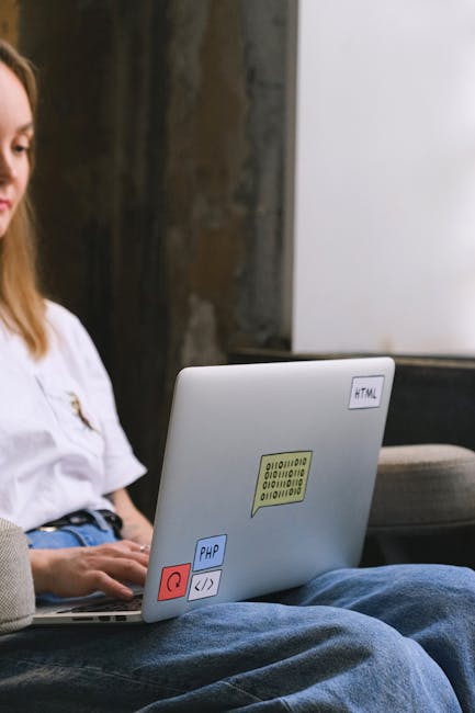 A silver laptop with a black keyboard is positioned on a wooden surface, displaying lines of programming code on its screen with a white background. The setting appears to be indoors, with a blurred b